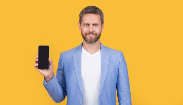 businessman showing portable phone. businessman presenting screen of portable phone with copy space. businessman with portable phone isolated on yellow. businessman hold portable phone in studio. - 写真・画像