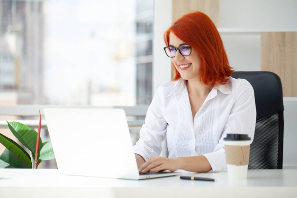 Beautiful smiling young red-haired business lady posing in office - Photo, Image