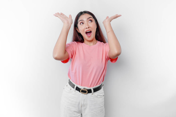 Mujer asiática conmocionada está usando camiseta rosa, señalando el espacio de copia en la parte superior de ella, aislado por el fondo blanco - Foto, imagen