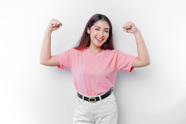 Excited Asian woman wearing a pink t-shirt showing strong gesture by lifting her arms and muscles smiling proudly - Foto, Imagen