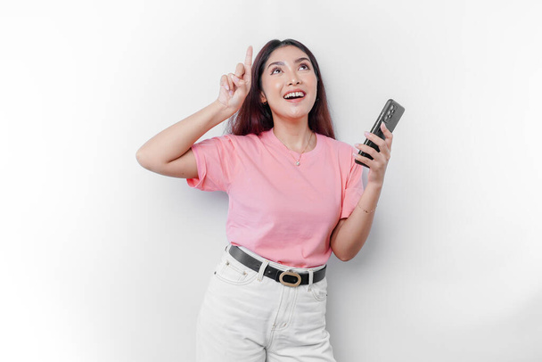 A portrait of a happy Asian woman wearing pink t-shirt, holding her phone, and pointing copy space on top of her, isolated by white background - Φωτογραφία, εικόνα