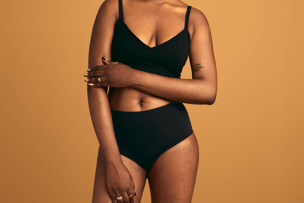 Anonymous African American plump female model in black underwear keeping hand on arm while standing against beige background in studio - Фото, изображение