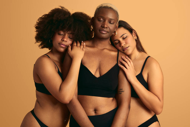 Tender multiethnic girlfriends in black underwear hugging gently against orange background concept of togetherness and support - Foto, afbeelding