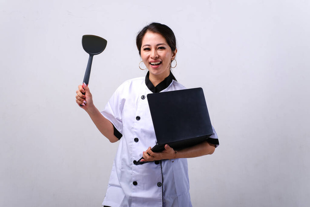 Smiling female Asian chef holding spatula and laptop isolated on white background - Valokuva, kuva