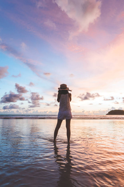 Vertical rear view of young adult traveler asian woman walking relax on beach sand with beautiful dramatic sunset sky. Travel on summer vacations. - Foto, Imagem