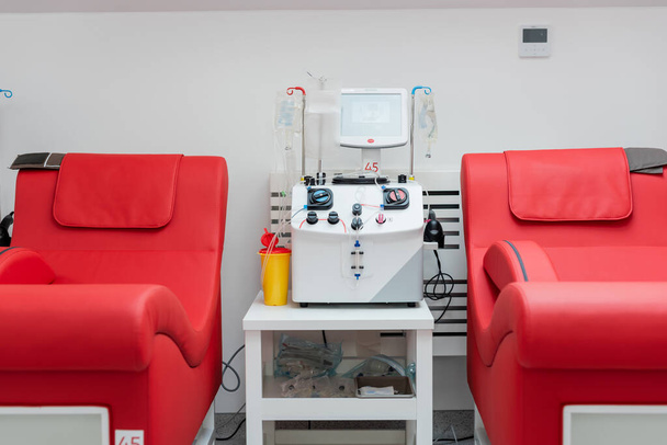 sillas médicas con diseño ergonómico cómodo cerca de la máquina de transfusión automatizada, pantalla táctil, taza de plástico y soportes de goteo con bolsas de infusión en el centro de donación de sangre - Foto, Imagen