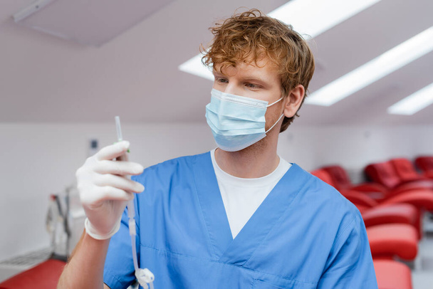 young and redhead doctor in blue uniform, medical mask and latex glove holding blood transfusion set near ergonomic medical chairs on blurred background in hospital - Photo, Image