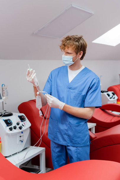 médico pelirrojo en uniforme azul, máscara médica y guantes de látex que sostienen el conjunto de transfusión cerca del equipo automatizado y cómodas sillas médicas en el centro de donación de sangre - Foto, Imagen