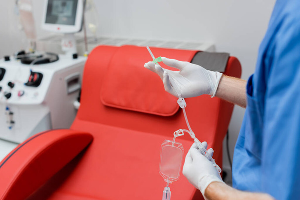 częściowy widok praktykującego w sterylnych rękawiczkach lateksowych z zestawem do transfuzji krwi w pobliżu krzesła medycznego o komfortowym ergonomicznym designie i nowoczesnym wyposażeniu na nieostrym tle w laboratorium - Zdjęcie, obraz
