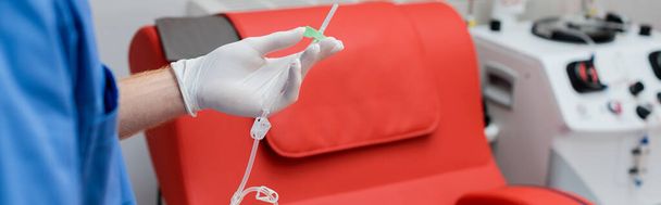 Ausgeschnittene Ansicht des Arztes in sterilen Latex-Handschuhen mit Transfusionsset in der Nähe verschwommener automatischer Geräte und medizinischer Stuhl im Blutspendezentrum, Banner - Foto, Bild