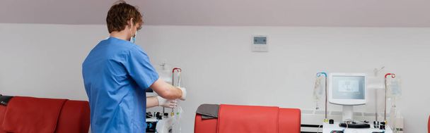 vista posterior del médico pelirrojo en uniforme azul, máscara médica y guantes de látex que trabajan con equipos de transfusión de sangre cerca de sillas médicas rojas y soportes de goteo en el laboratorio médico, pancarta - Foto, Imagen