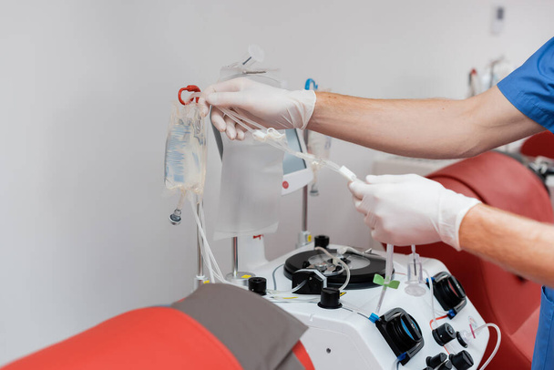 vista parcial del médico en guantes de látex estériles ensamblando transfusión de sangre en soporte de goteo con bolsas de perfusión cerca de equipos automatizados y sillas médicas rojas en laboratorio médico  - Foto, Imagen