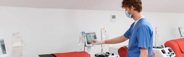 vista lateral del médico en máscara médica y uniforme azul de pie cerca de la pantalla táctil y la operación de la máquina de transfusión de sangre en el laboratorio médico estéril, pancarta - Foto, Imagen