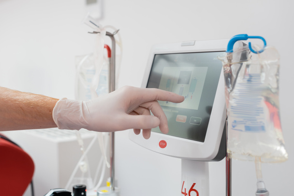 vista parcial del trabajador sanitario en guante de látex operando una moderna máquina de transfusión automatizada con pantalla táctil cerca de soportes de goteo y bolsas de infusión en laboratorio médico - Foto, Imagen