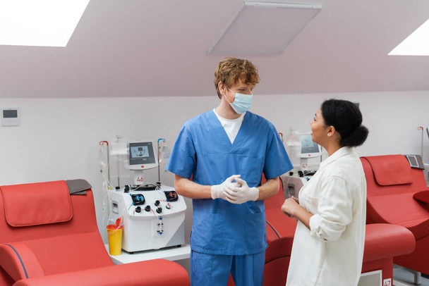 πολυφυλετική γυναίκα και κοκκινομάλλα γιατρός με μπλε στολή, ιατρική μάσκα και γάντια λατέξ μιλάμε κοντά σε ιατρικές καρέκλες και αυτοματοποιημένη μηχανή μετάγγισης στο κέντρο αιμοδοσίας - Φωτογραφία, εικόνα