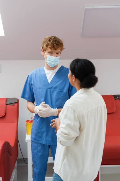 mladý a zrzavý lékař v lékařské masce, latexové rukavice a modrá uniforma mluví s multiraciální pacient v blízkosti červených lékařských židlí v transfuzní stanici - Fotografie, Obrázek