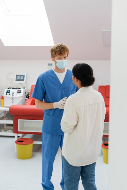 redhead doctor in medical mask, blue uniform and latex gloves talking to multiracial near medical chairs and automated transfusion machine on blurred background - Photo, Image