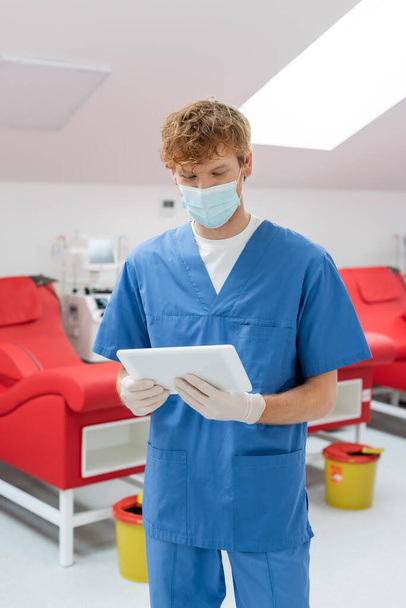 рудий лікар в медичній масці, блакитна уніформа і латексні рукавички дивиться на цифровий планшет біля медичних крісел і машини переливання в центрі донорства крові на розмитому фоні
 - Фото, зображення
