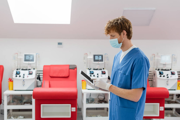 médico pelirrojo en máscara médica, uniforme azul y guantes de látex usando tableta digital con pantalla en blanco cerca de sillas médicas borrosas y máquinas automatizadas de transfusión en el centro de donación de sangre - Foto, Imagen
