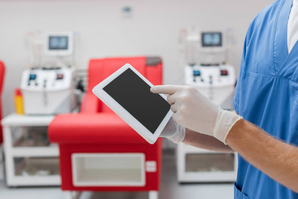 részleges kilátás orvos latex kesztyű mutató digitális tabletta üres képernyőn közelében elmosódott orvosi szék és vérátömlesztő gép elmosódott háttér klinikán - Fotó, kép