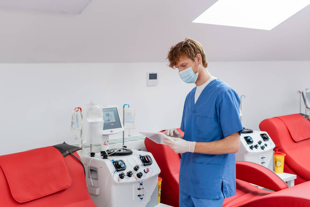 médico pelirrojo en uniforme azul, máscara médica y guantes de látex mirando la tableta digital cerca de sillas médicas y máquinas automatizadas de transfusión en el centro de donación de sangre - Foto, Imagen