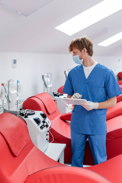 vöröshajú orvos orvosi maszkban digitális tablettát tart, és nézi csepegtető standok közelében ergonomikus székek és automatizált transzfúziós gépek véradó központ - Fotó, kép