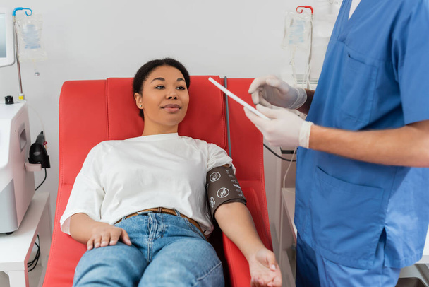multiracionális nő a vérnyomás mandzsettát ül orvosi széken közelében automatizált transzfúziós gépek és nézi orvos kék egyenruhában és latex kesztyű segítségével digitális tabletta a klinikán  - Fotó, kép