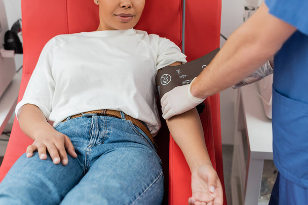 Doktorun lateks eldivenli kısmi görüntüsü. Kan nakli istasyonunda rahat bir tıbbi sandalyede oturan çok ırklı kadına baskı kelepçesi takıyor. - Fotoğraf, Görsel