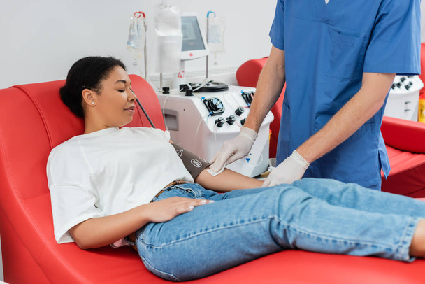 kék egyenruhás egészségügyi dolgozó és latex kesztyű alkoholos törlőkendőt és vérnyomásmérőt tartó multiracionális nő karját tartva az orvosi széken ülve a transzfúziós gép közelében - Fotó, kép