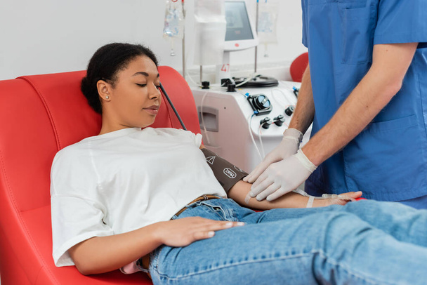 Arzt in Latex-Handschuhen klebt Pflaster am Arm einer multiethnischen Frau, die auf dem Behandlungsstuhl sitzt und in der Nähe eines automatischen Transfusionsgeräts Blut spendet - Foto, Bild