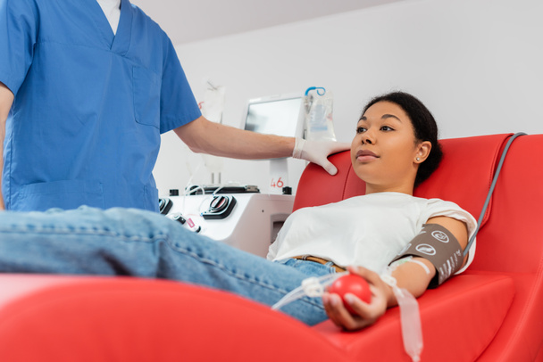 arts in blauw uniform staan in de buurt van multiraciale vrouw zitten op comfortabele medische stoel in de buurt van transfusie machine terwijl het doneren van bloed in medisch laboratorium - Foto, afbeelding