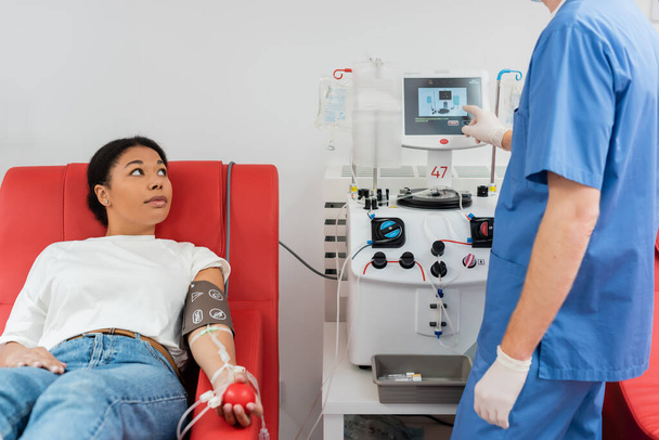 Врач в синей униформе и латексных перчатках операционный аппарат для переливания крови рядом с многорасовой женщиной, сидящей на медицинском стуле с резиновым мячом во время сдачи крови в лаборатории - Фото, изображение