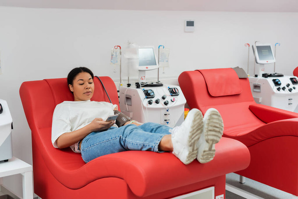 mujer multirracial navegar por Internet en el teléfono inteligente mientras está sentado en la silla médica cómoda cerca de la máquina de transfusión, mientras que la donación de sangre en la clínica, procedimiento médico - Foto, imagen