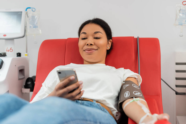 potěšen multiraciální žena v krevní tlak manžety zprávy na mobilním telefonu, zatímco sedí na lékařské židli v blízkosti automatizovaného transfuzního stroje v moderní laboratoři, lékařský zákrok  - Fotografie, Obrázek