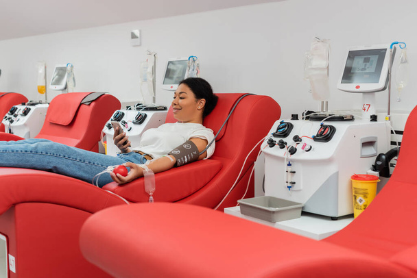 sorridente donna multirazziale con palla di gomma e trasfusione set navigazione internet su smartphone mentre seduto sulla sedia medica vicino attrezzature automatizzate durante la donazione di sangue in clinica - Foto, immagini