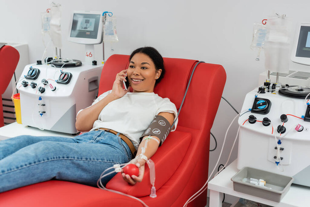 счастливая многорасовая женщина с резиновым мячом сидит на медицинском стуле и разговаривает по мобильному телефону возле аппаратов для переливания крови во время сдачи крови в лаборатории - Фото, изображение