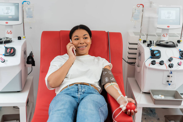 mujer multirracial sonriente con manguito de presión y bola de goma sentado en la silla médica cerca de las máquinas de transfusión y hablando por teléfono móvil durante la donación de sangre en la clínica - Foto, Imagen