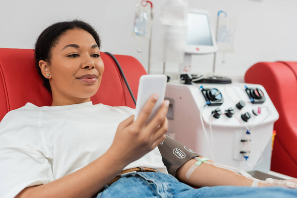 elégedett multiraciális nő ül kényelmes orvosi szék és üzenetküldés mobiltelefon közelében automatizált vérátömlesztő gép laboratóriumi, homályos háttér - Fotó, kép