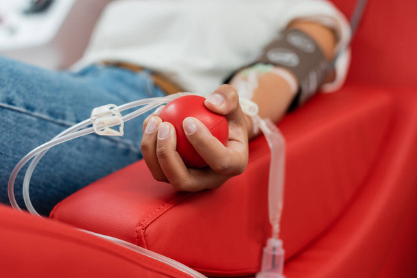 enfoque selectivo de la bola de goma en la mano de la mujer multirracial con transfusión conjunto sentado en la silla médica cómoda y la donación de sangre en el laboratorio - Foto, Imagen