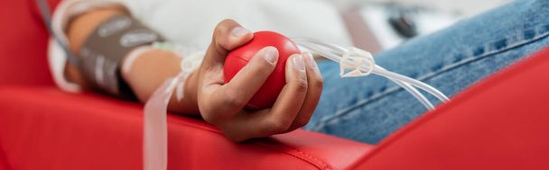 vista parcial de la mujer multirracial con el sistema de transfusión que sostiene la bola de goma mientras está sentado en la silla médica ergonómica durante la donación de sangre en el laboratorio, pancarta - Foto, Imagen