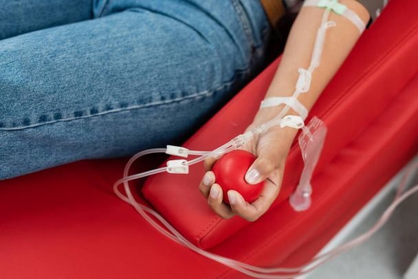 vista parcial de la mujer multirracial con un conjunto de transfusión que sostiene la bola de goma mientras está sentado en la silla médica ergonómica durante la donación de sangre en la clínica, procedimiento médico  - Foto, Imagen