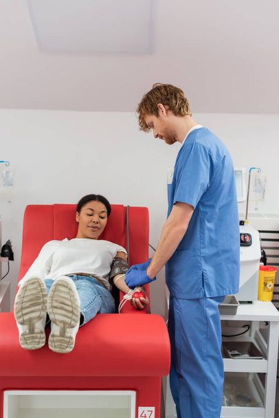 vörös hajú, kék egyenruhás egészségügyi dolgozó, aki a transzfúziót az orvosi székben gumilabdával a kezében ülő multiracionális nőn állítja be, miközben vért ad a klinikán - Fotó, kép