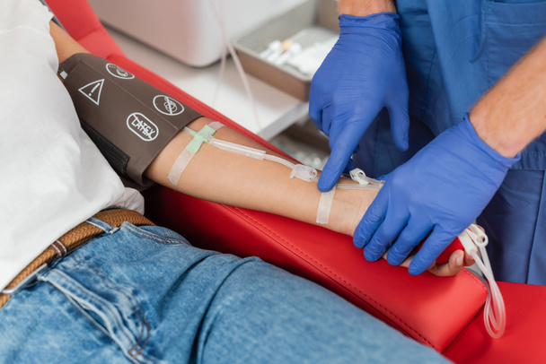 vista recortada del médico en guantes de látex pegando una tirita en el brazo de una mujer multirracial con manguito de presión y transfusión durante la donación de sangre en la clínica - Foto, imagen