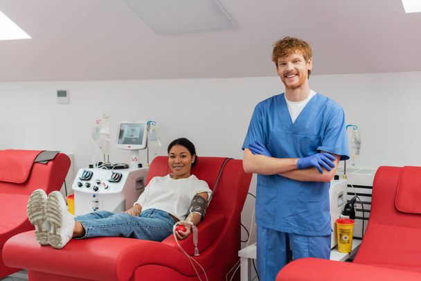 happy redhead doctor in blue uniform and latex gloves looking at camera near transfusion machines and multiracial woman on medical chair donating blood in hospital - Photo, Image