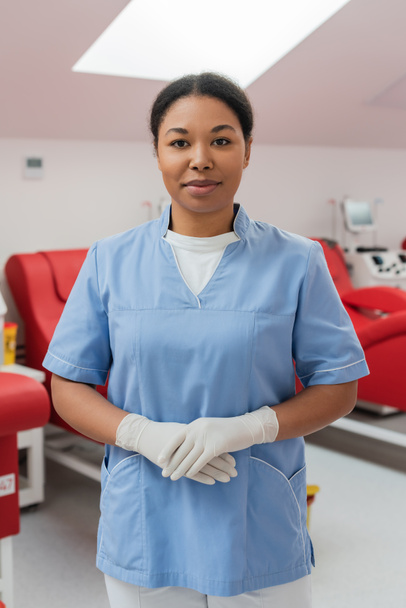 positive multiracial nurse in blue uniform and latex gloves looking at camera near medical chairs and transfusion machines in blood donation center on blurred background - Photo, Image