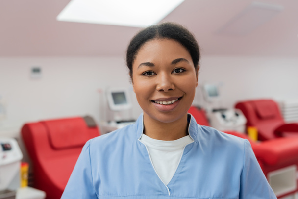 cheerful multiracial nurse in blue uniform smiling and looking at camera near medical chairs and transfusion machines on blurred background in blood donation center - Photo, Image