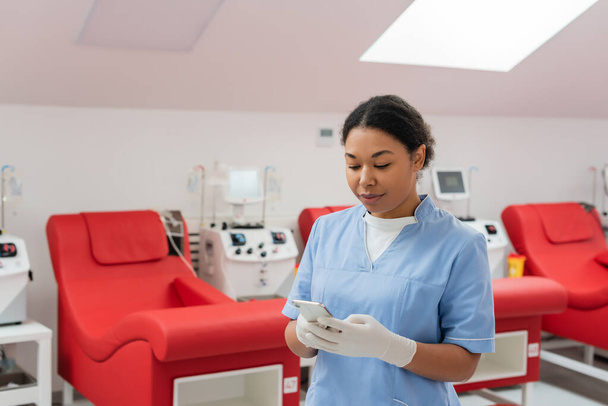 multiraciale verpleegkundige in blauw uniform en latex handschoenen messaging op mobiele telefoon in de buurt van medische stoelen en transfusie machines in bloed donatie centrum - Foto, afbeelding