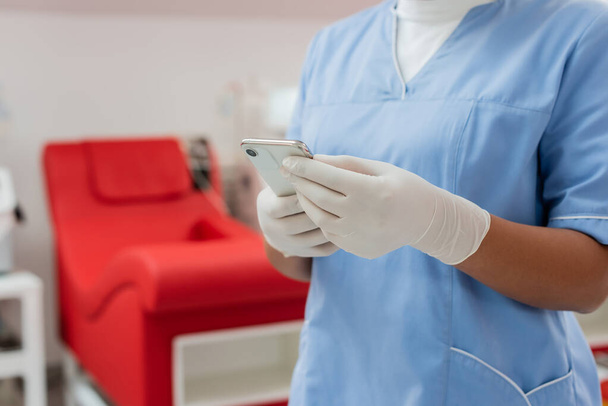 vista parcial de la enfermera multirracial en uniforme azul y guantes de látex estériles mensajería en el teléfono móvil cerca de la silla médica borrosa en el centro de transfusión de sangre  - Foto, imagen