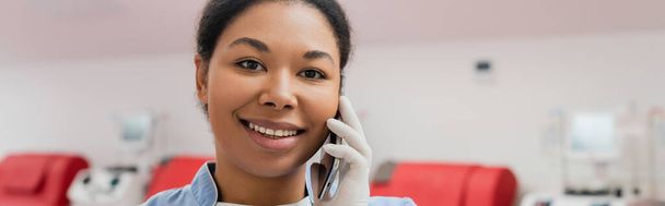 joven y feliz trabajador de la salud multirracial en guante de látex estéril sonriendo durante la conversación en el teléfono móvil en la estación de transfusión de sangre, pancarta - Foto, Imagen