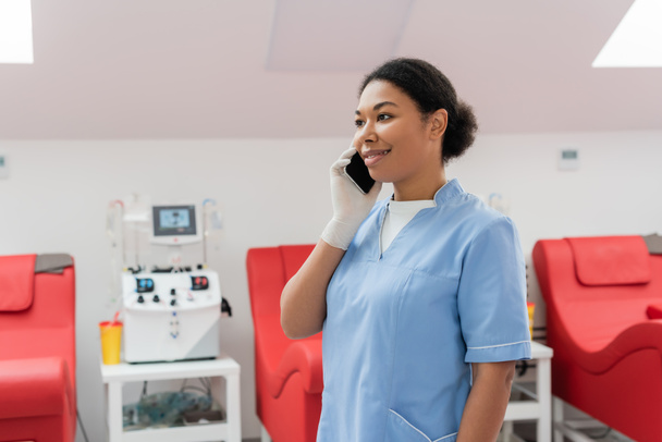 szczęśliwy wielorasowy pracownik służby zdrowia w niebieskim mundurze i rękawicy lateksowej rozmawiający przez telefon komórkowy w pobliżu rozmytej maszyny transfuzyjnej i krzeseł medycznych w centrum krwiodawstwa - Zdjęcie, obraz